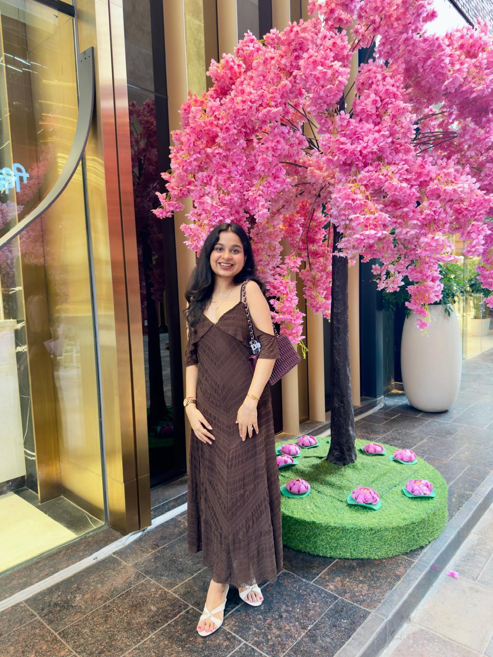Woman in a brown dress standing and smiling in front of a vibrant pink flowering tree outside a modern storefront
